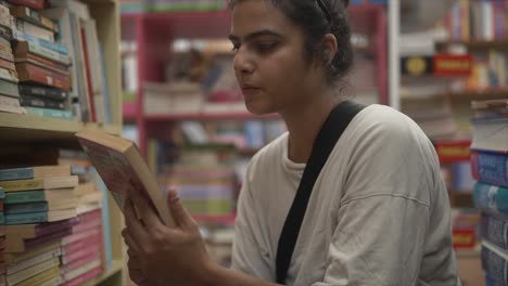 Asian-girl-sitting-and-exploring-books-through-bookshelves,-Side-angle-close-up-shot