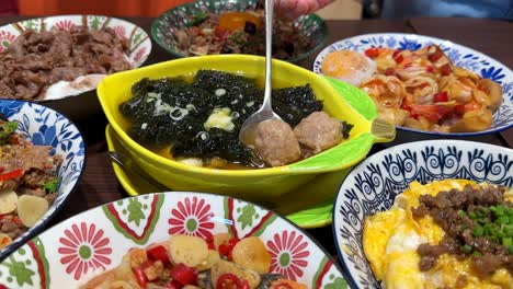 a hand uses a spoon to serve seaweed soup with meatballs from a yellow bowl, surrounded by vibrant asian dishes under bright indoor lighting