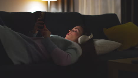 Mujer-Con-Auriculares-Inalámbricos-Recostada-En-Un-Sofá-En-Casa-Por-La-Noche-Transmitiendo-O-Mirando-Contenido-En-Línea-En-Un-Teléfono-Móvil-3