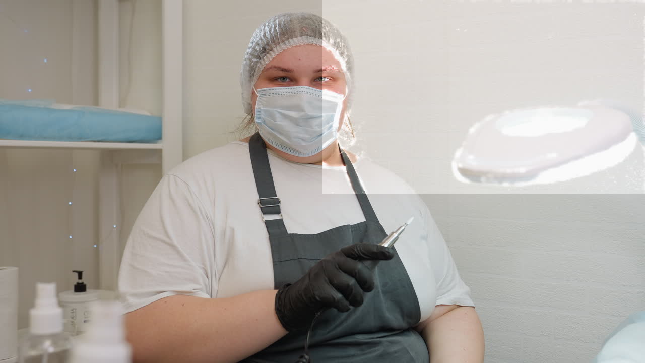 Technician wearing face mask and disposable shower cap holds electric file while seated under bright salon lamp. Dressed in white shirt and dark apron, she is ready for professional nail care session