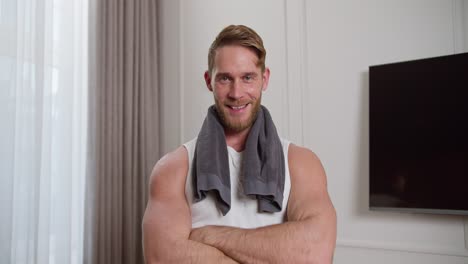 Portrait-of-a-confident-blond-man-with-stubble-in-a-white-T-shirt-with-an-athletic-build-who-holds-a-gray-towel-on-his-shoulders-and-neck-and-looks-at-the-camera-after-playing-sports-and-training-in-a-modern-apartment-in-the-morning