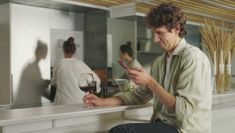 man using smartphone and drinking wine in a modern kitchen with a woman in the background