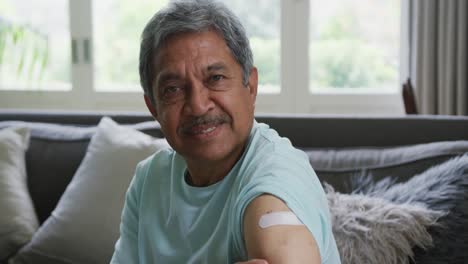 portrait of senior man touching bandage on arm and smiling while sitting at home after vaccination