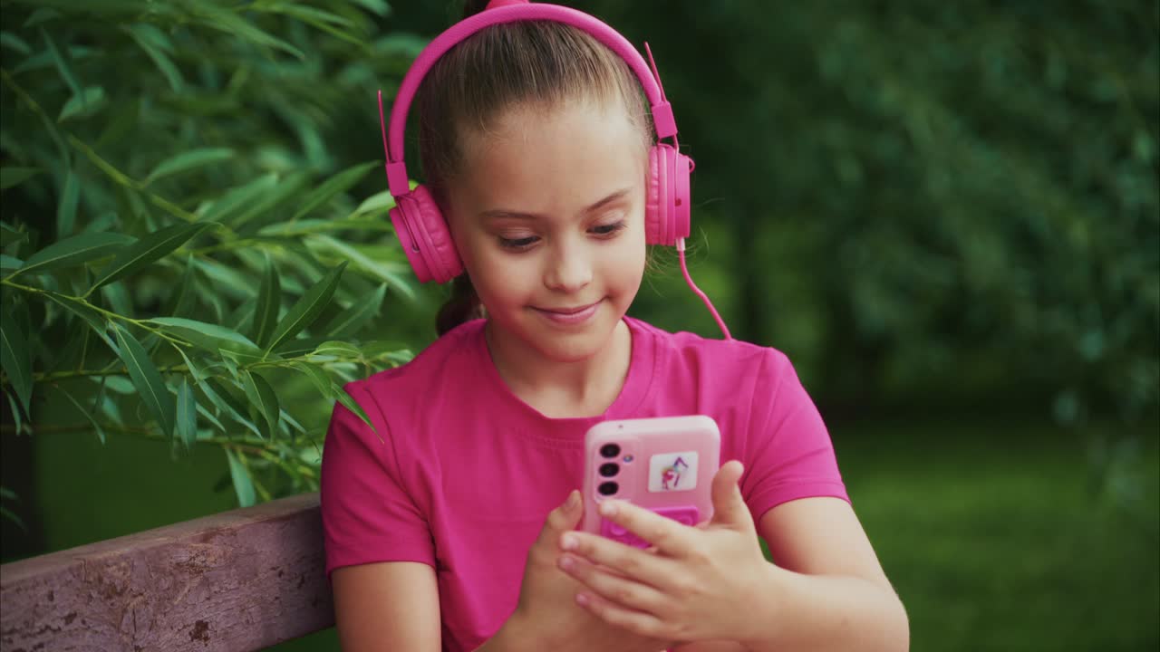 A Young Girl in Pink Headphones Enjoys Her Smartphone while Relaxing on a Bench Surrounded by Lush Greenery, Capturing a Moment of Joy and Connection with Music
