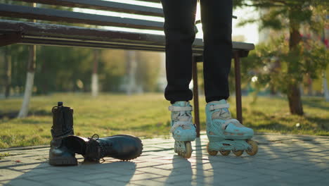 a person dressed in black trousers is standing near a park bench, ready to skate with rollerblades on their feet. a pair of black boots is placed on the ground beside them