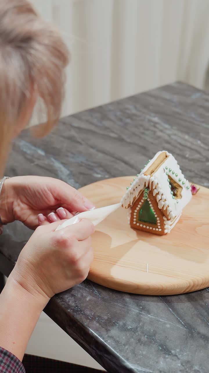 vista aérea de una mujer decorando un pastel en forma de casa con intrincados detalles de glaseado en una bandeja de madera, flores secas rodean el espacio de trabajo, con una silla visible en un fondo borroso