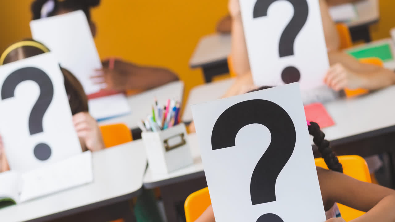 Diverse class of elementary schoolchildren at their desks holding question mark signs