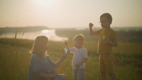 a happy woman in a blue dress kneels in a grassy field during sunset, holding a bubble bottle while her younger son dips a wand and blows bubbles. her older son is also blowing bubbles