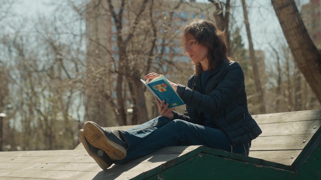 Woman reads book while sitting on wooden playground equipment in outdoor park setting with trees and buildings visible. Woman wears casual clothing including jeans and sneakers as woman
