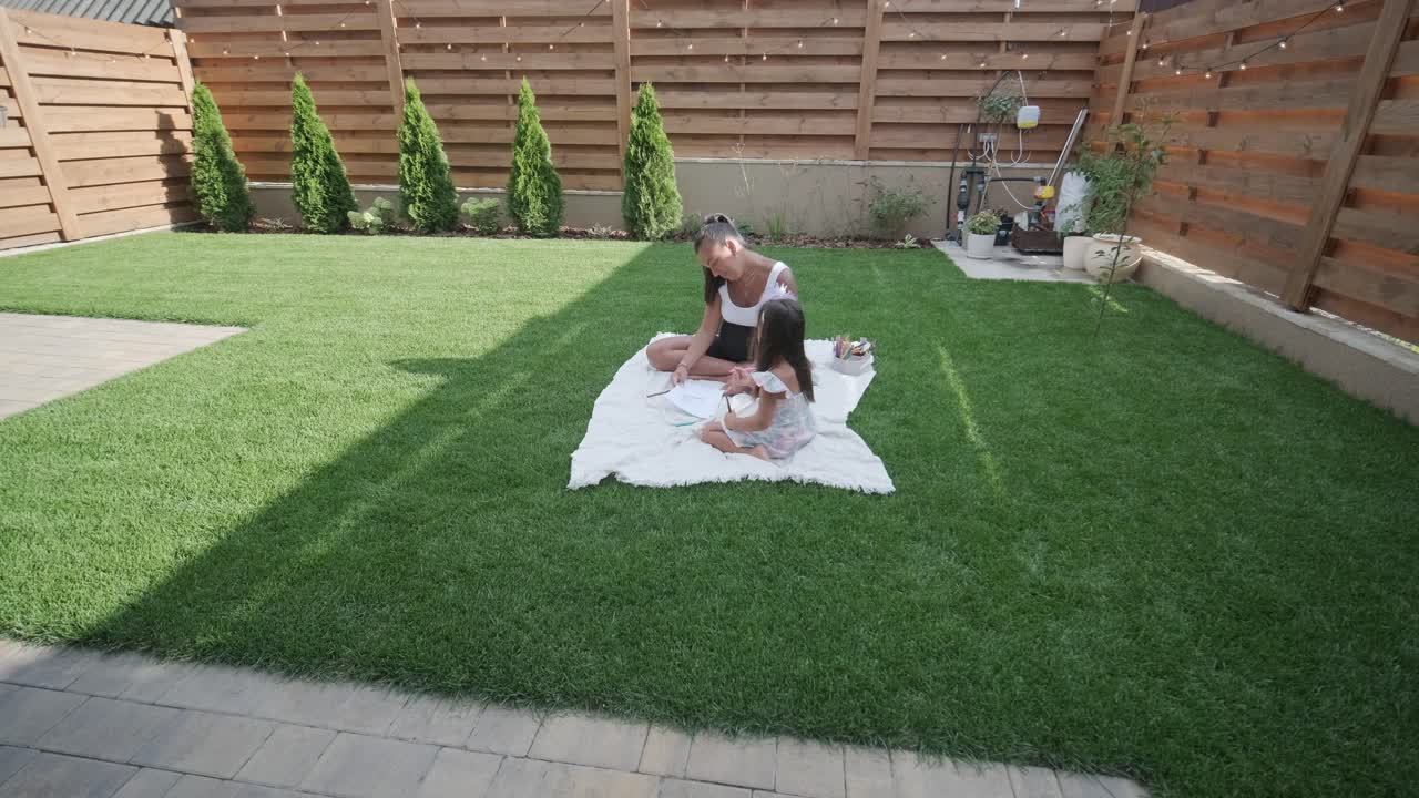 A mother and her young daughter sit on a blanket in the backyard, engaging in a drawing activity together. The outdoor scene captures a peaceful family moment, fostering creativity and bonding.