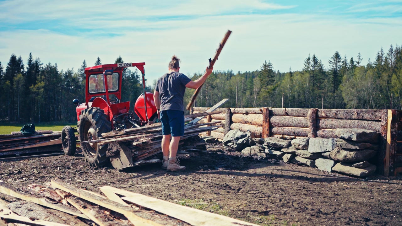 Man Carries Firewood Logs From A Tractor To Stack Them Beside A Wooden Fence On A Farmstead. - wide shot