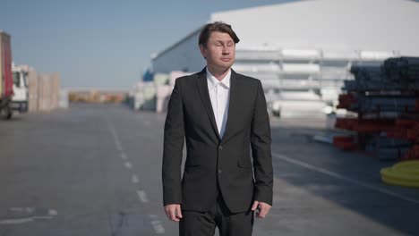 portrait of successful happy caucasian manufacturing business owner in suit raising hands looking around in slow motion standing at storage outdoors. positive wealthy man on sunny day at warehouse.