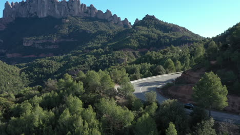 Cinematic-aerial-shot-of-old-black-car-entering-turn-on-mountain-road-with-montserrat-mountain-in-background