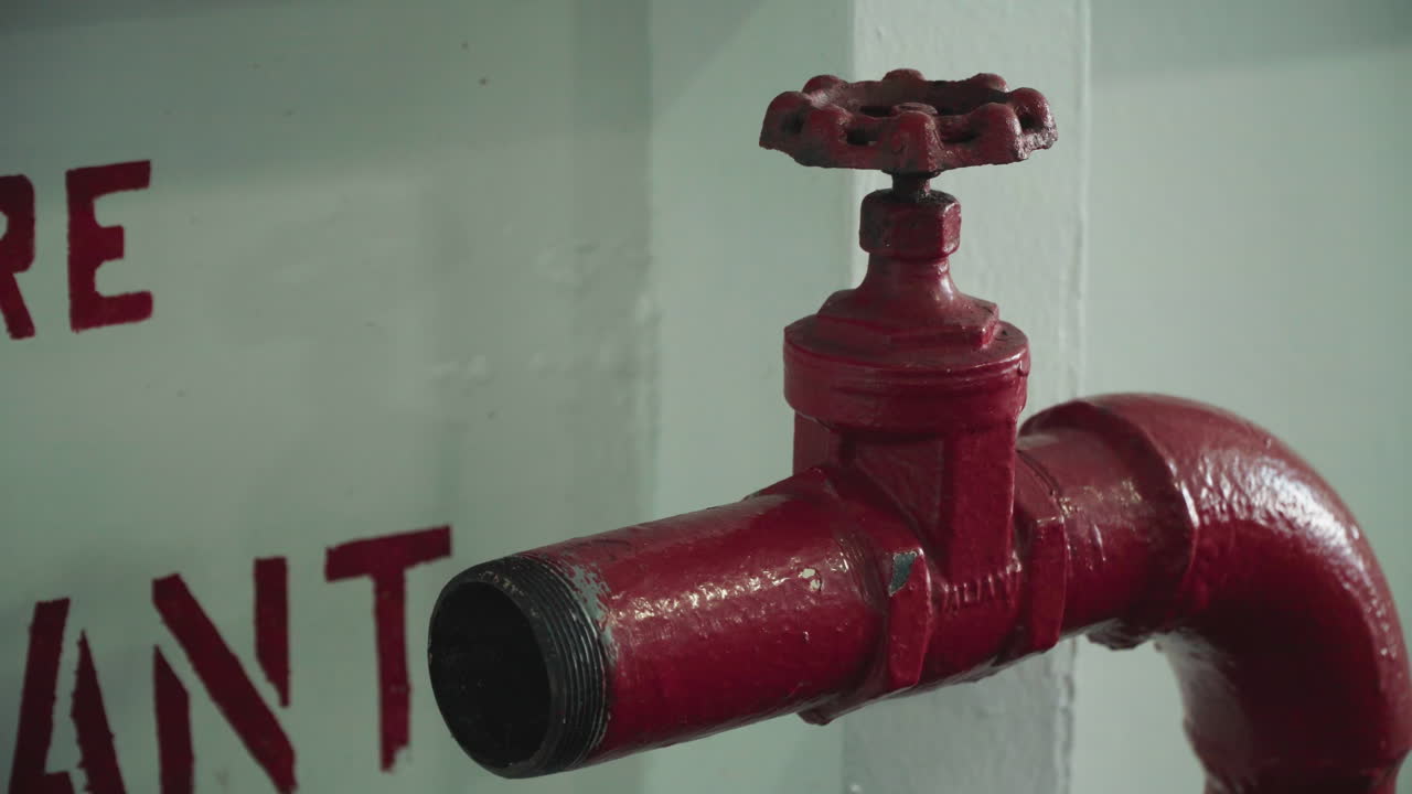 Valvola antincendio rossa montata a parete, primo piano che mostra il tubo verniciato lucido e il beccuccio filettato, segnaletica di emergenza parziale sullo sfondo, illuminazione fioca del corridoio, metallo testurizzato con usura e tracce.