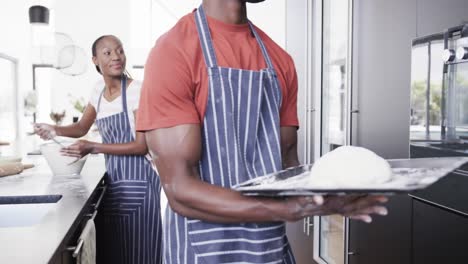 happy african american couple in aprons baking bread putting dough into oven in kitchen, slow motion