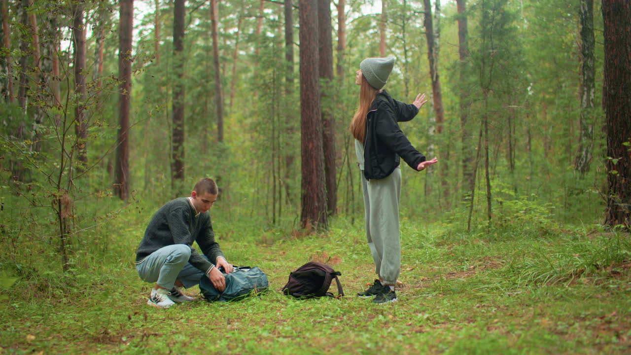 traveler in forest as man kneels to unzip backpack on grassy ground while woman stands nearby stretching and gazing upward thoughtfully, surrounded by tall trees and dense woodland atmosphere
