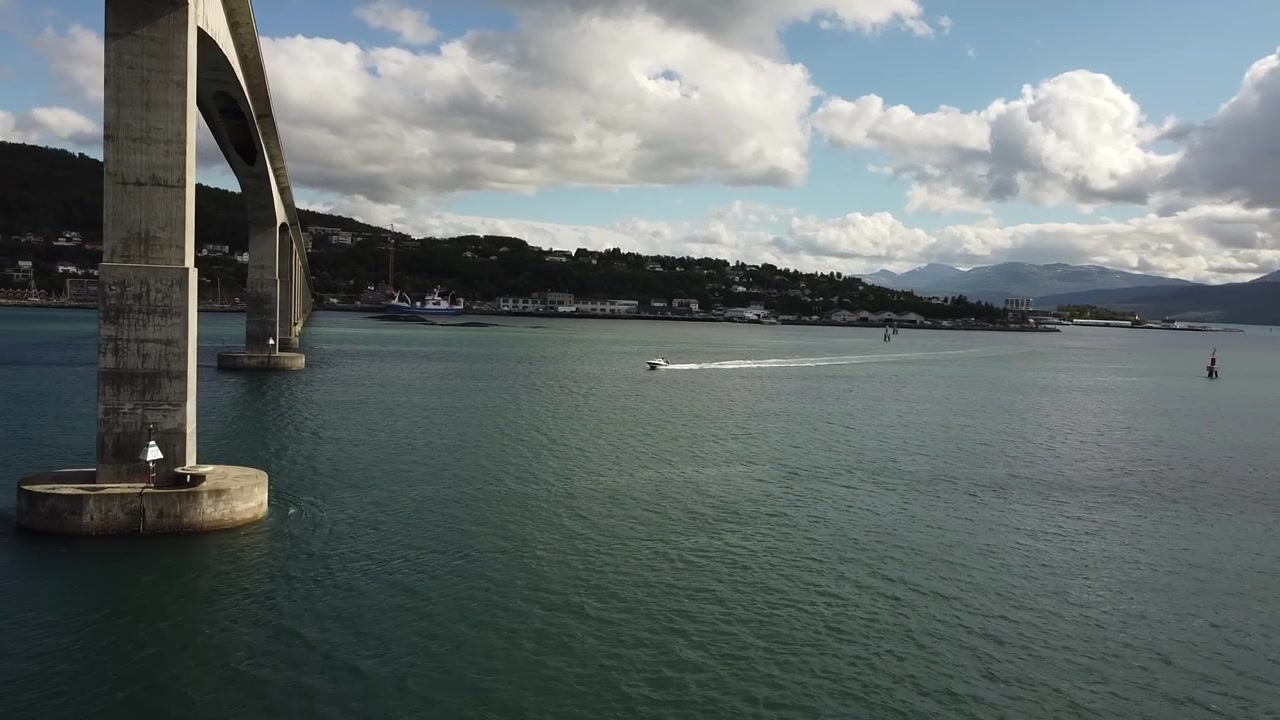 Imagens aéreas de drone orbitam para a esquerda sob a Ponte Gisund em Senja, Noruega, enquanto uma lancha se aproxima do fiorde aberto sob montanhas e nuvens dispersas