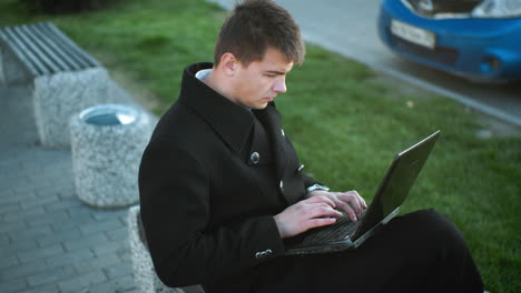 real estate mogul wearing black suit with silver buttons focused while typing on laptop outdoors, background features green grass, pavement, and blurred blue car parked nearby