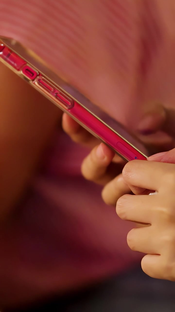 Primo piano della mano di una persona che scorre su uno smartphone con una custodia rossa, seduta al chiuso sotto un'illuminazione calda, con un movimento fluido della fotocamera e una profondità di campo ridotta.