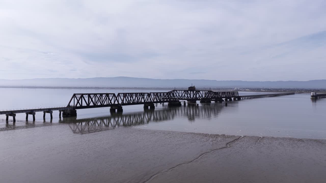 Drone che vola lentamente all'indietro mostrando una vista aerea dello storico e abbandonato Ponte ferroviario di Dumbarton che attraversa la Baia di San Francisco in una giornata nuvolosa e cupa, con montagne sullo sfondo