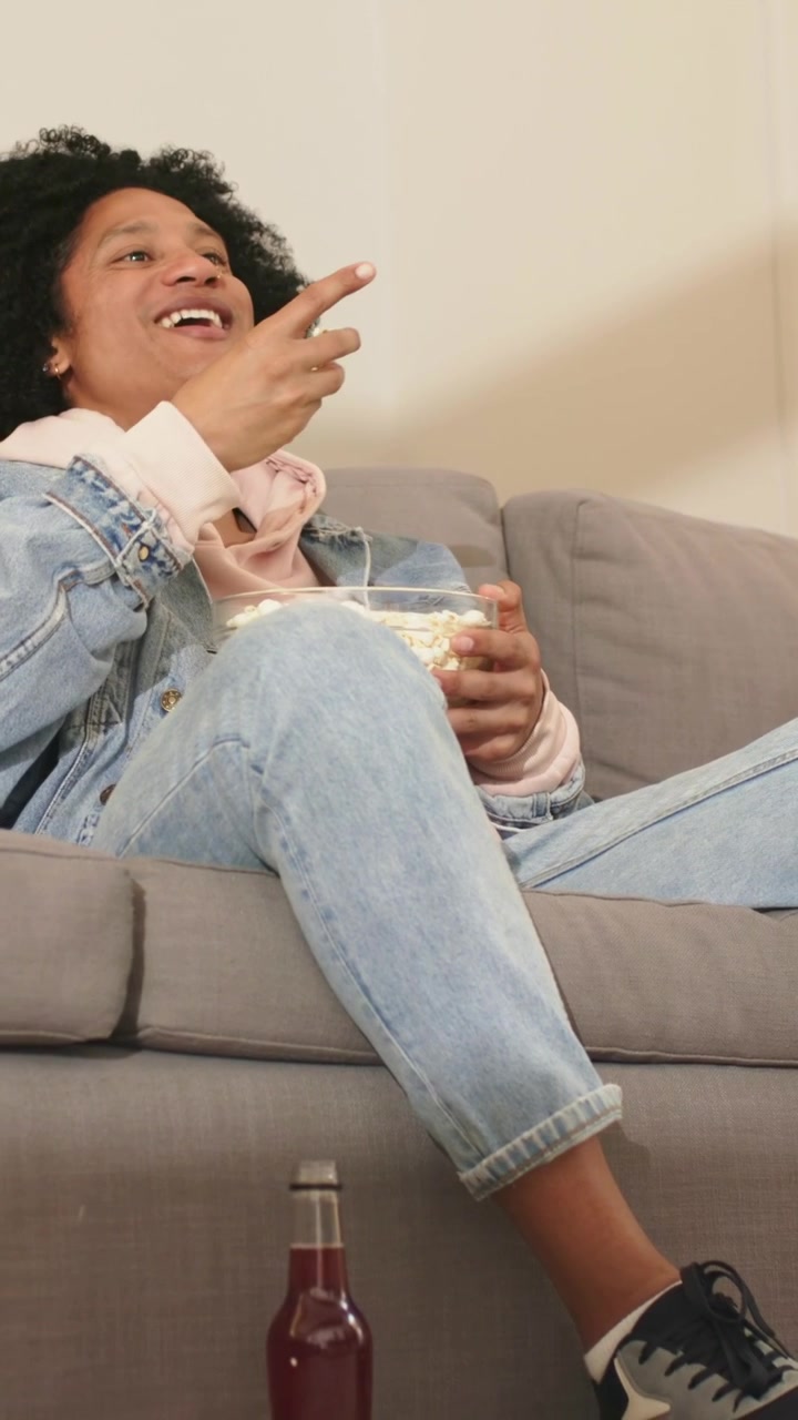 Vertikal video: Mand i denim trykker på fjernbetjening, mens et show starter, ser tv og spiser popcorn derhjemme.