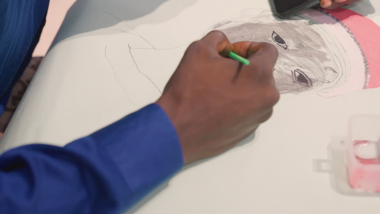 Closeup black artist hand sketching portrait, green pencil tracing charcoal tones on white paper, blue shirt sleeve visible, smartphone reference nearby, small paint box at edge, intimate focused