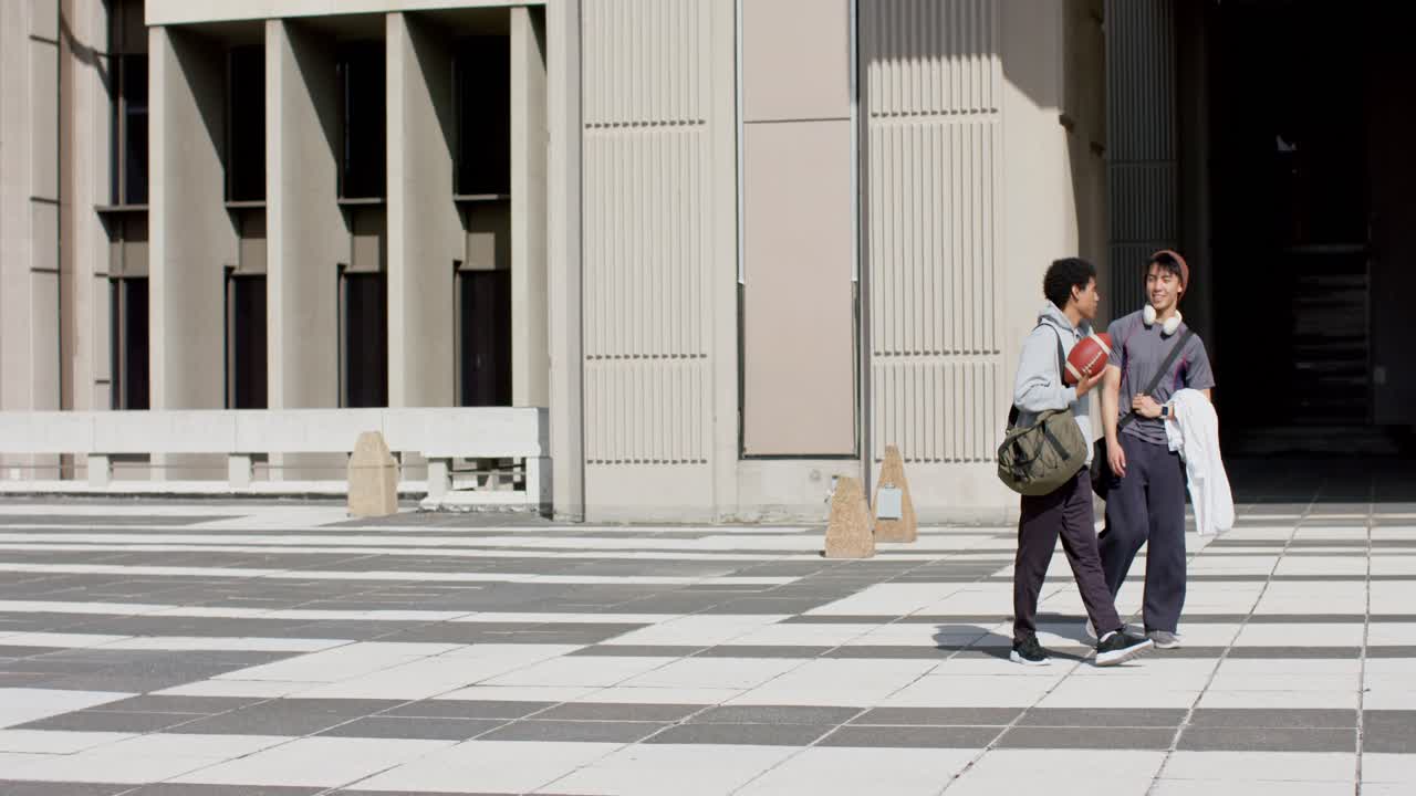 Dopo aver chiacchierato, amici uomini di diverse etnie camminano nella piazza del campus con un pallone da football e una borsa, con spazio per il testo.