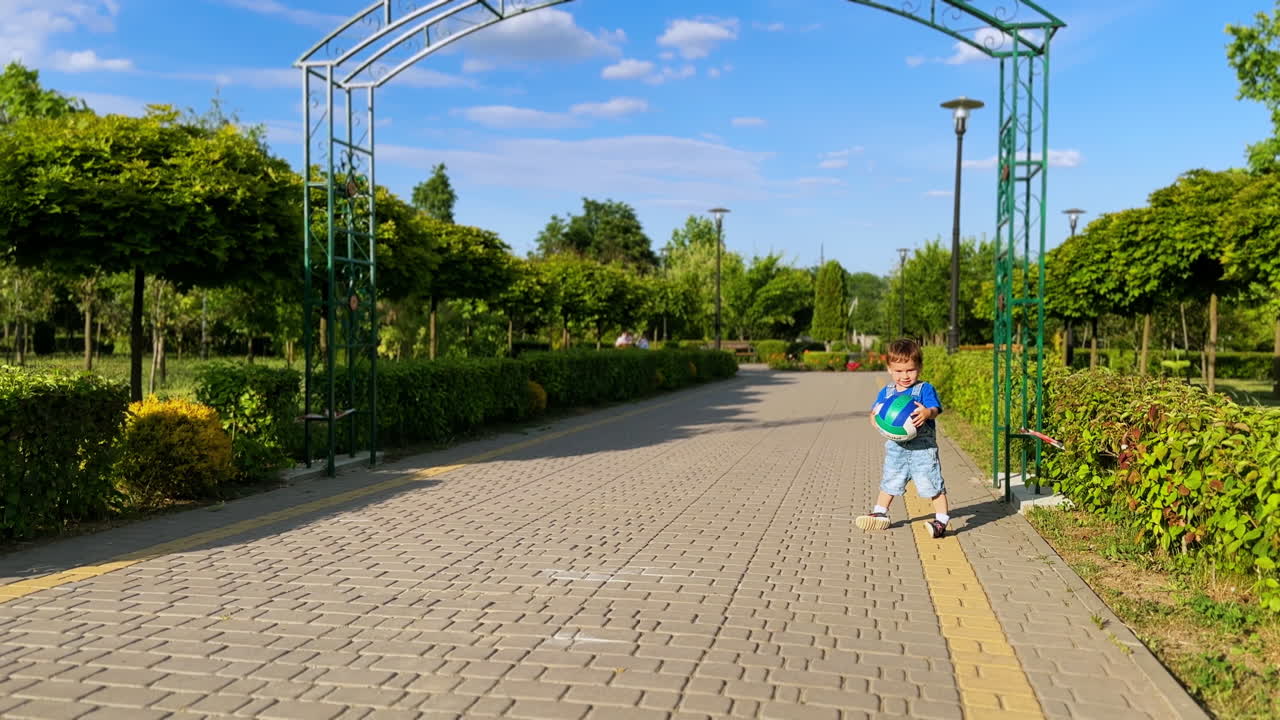 Caucasian baby boy carrying a ball in the park. Adorable kid stops on the paved alley in the park.