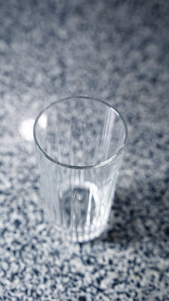 Long ice cube is placed into the glass with metal tongs. Top view on the glass standing on the table. Beverage preparation. Vertical video.