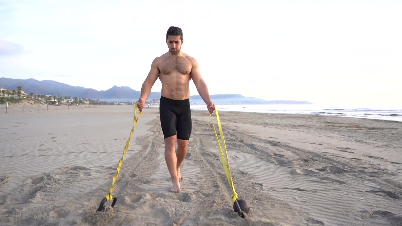 entrenamiento metabólico en la playa con un hombre musculoso que realiza una variante de pecho y goma con pesas rusas al amanecer