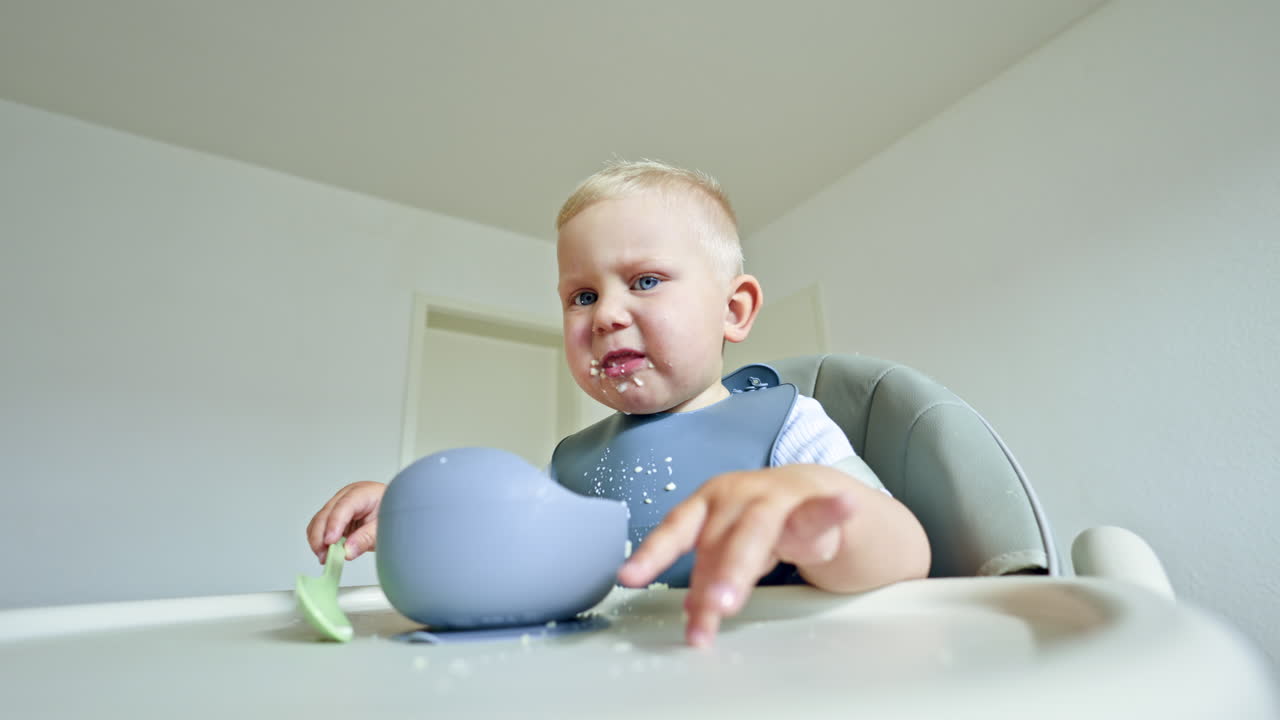 A little boy with a silicone apron sits in a high chair and eats porridge with a spoon on his own. Toddler eats breakfast on his own and chews happily. Safe silicone utensils for children
