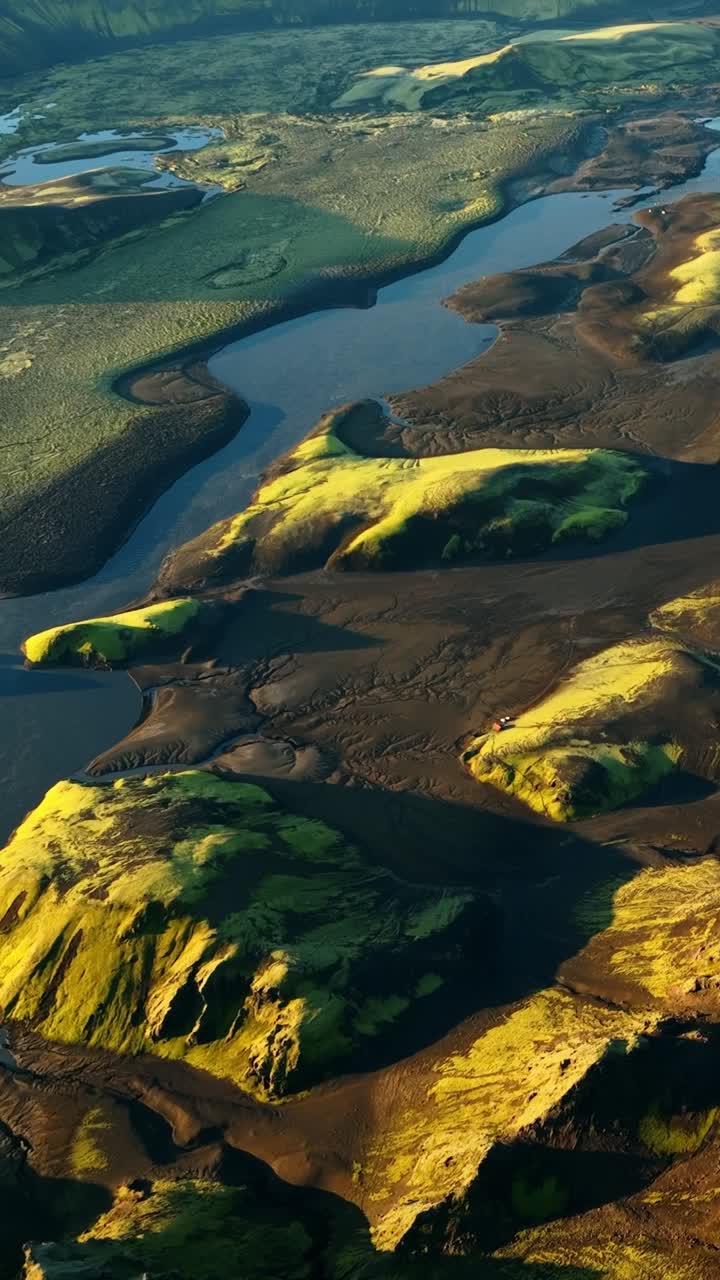 Vista aérea vertical con dron de las tierras altas volcánicas islandesas con colinas cubiertas de musgo, llanuras de arena negra y un río azul serpenteante durante la hora dorada.