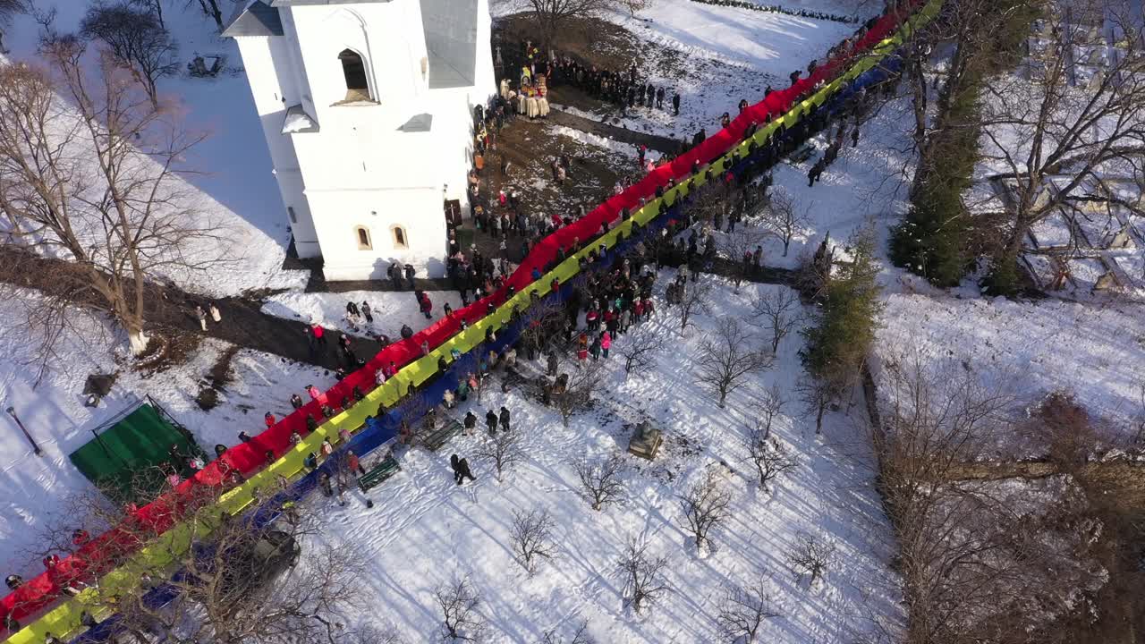 Static aerial close-up of a large crowd holding a massive, extended Romanian flag, tricolor on the snow-covered grounds of Raducanu Monastery in Targu Ocna during a winter national celebration