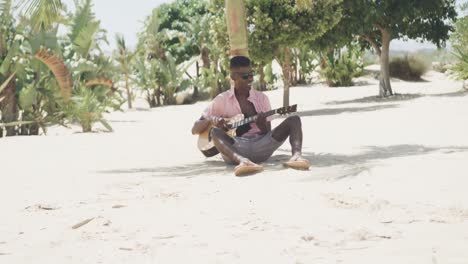 Feliz-Hombre-Afroamericano-Sentado-Bajo-Un-árbol-Tocando-La-Guitarra-Y-Cantando-En-La-Playa-Soleada,-Cámara-Lenta
