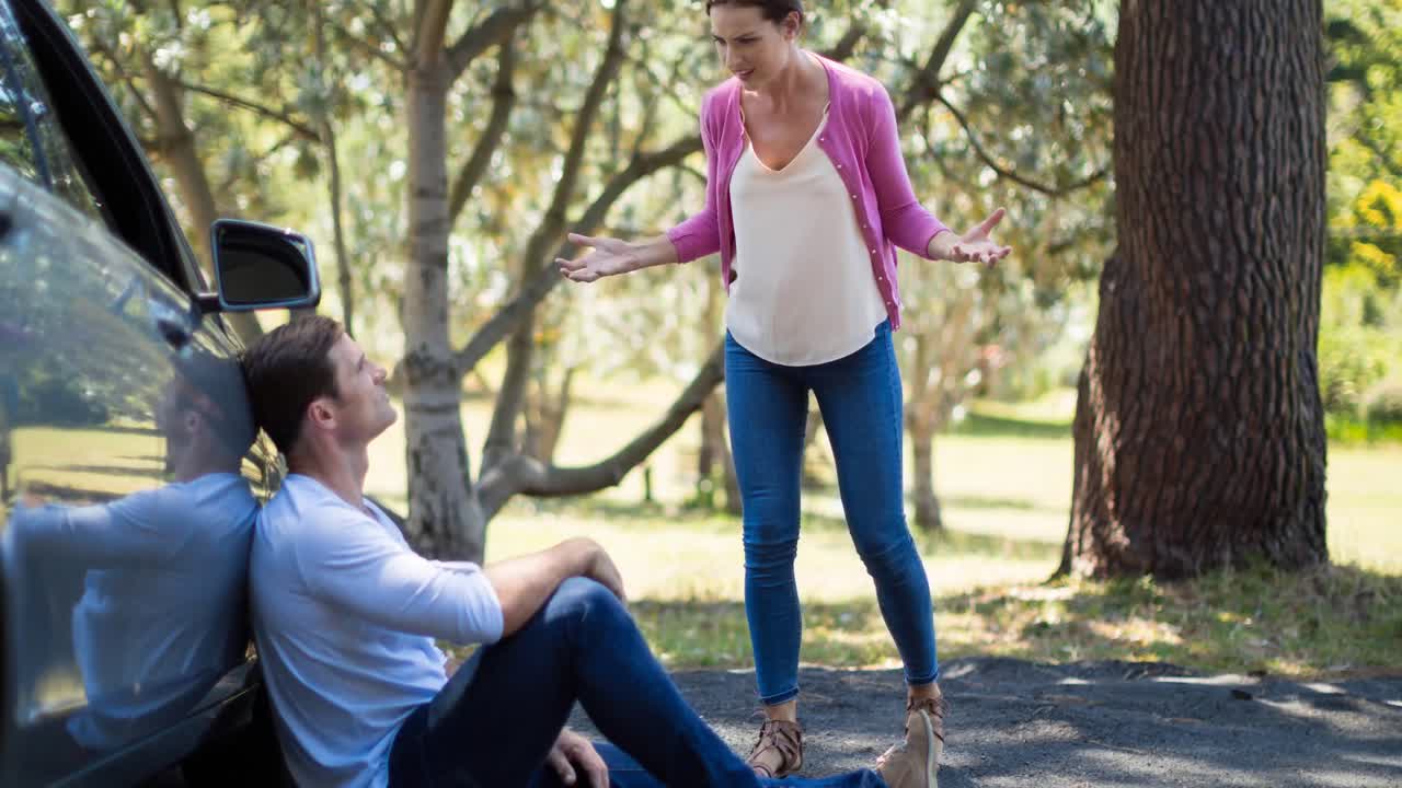 mujer caucásica enojada discutiendo con su compañero masculino, sentada en la carretera al lado de un coche averiado