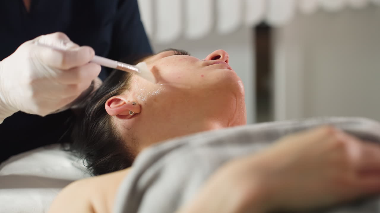 Female client lies with eyes closed on spa bed while beauty expert wearing protective gloves dips brush into clear gel and smooth applies nourishing facial mask in gentle strokes for radiant skin