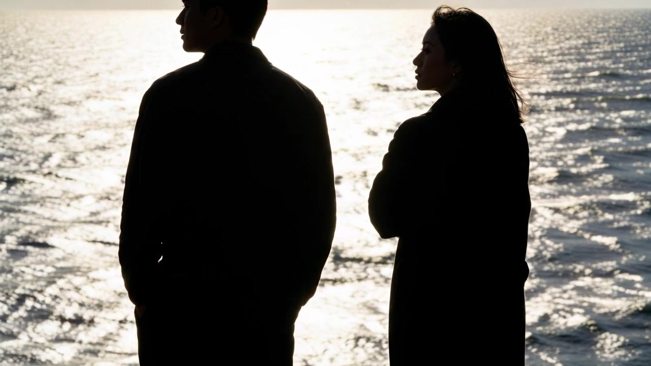 Standing silhouetted couple wearing dark long coats, watching sunset over water with sun reflection