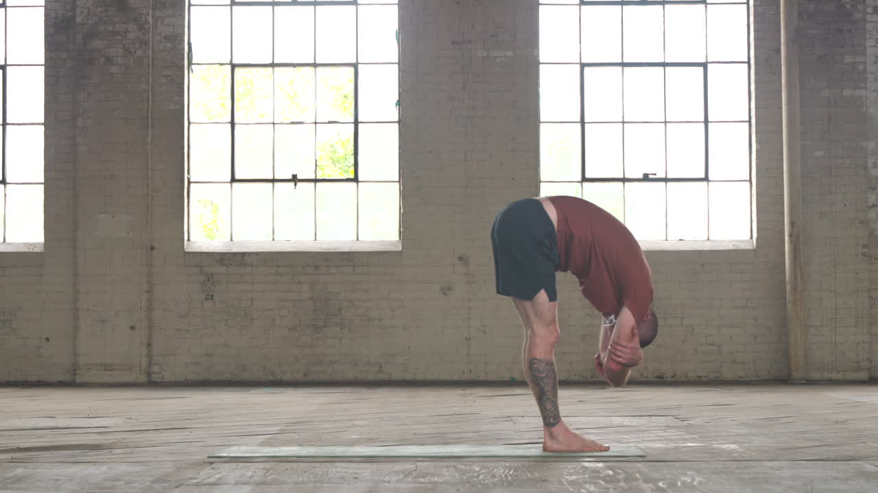 Man in an industrial warehouse practicing yoga, the pose of forward fold.