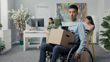 disabled ambitious man in shirt with asian korean beauty sits in wheelchair at desk in the company on knees holding cardboard box with packed things, gets fired from job unable to fulfill his duties