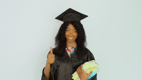una joven afroamericana con una túnica negra y un sombrero de maestro se mantiene erguida sosteniendo libros de texto y muestra un buen gesto. mira a la cámara. retrato de media altura