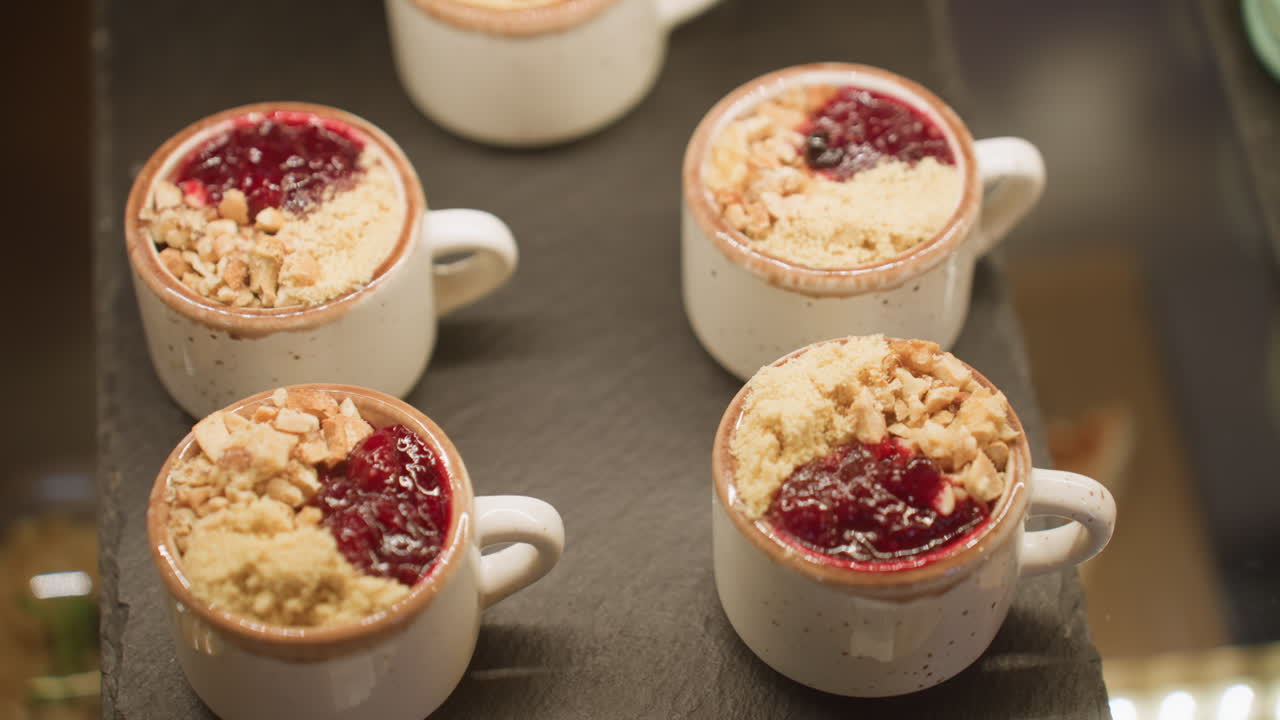 Close up of white ceramic dessert cups filled with red berry sauce, crushed nuts, and crumbs arranged neatly on black tray inside store display case under warm light