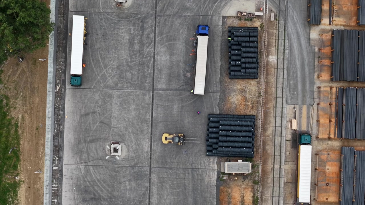 An aerial view shows trucks and a skid steer loader moving steel materials in a busy industrial factory yard in Hungary.