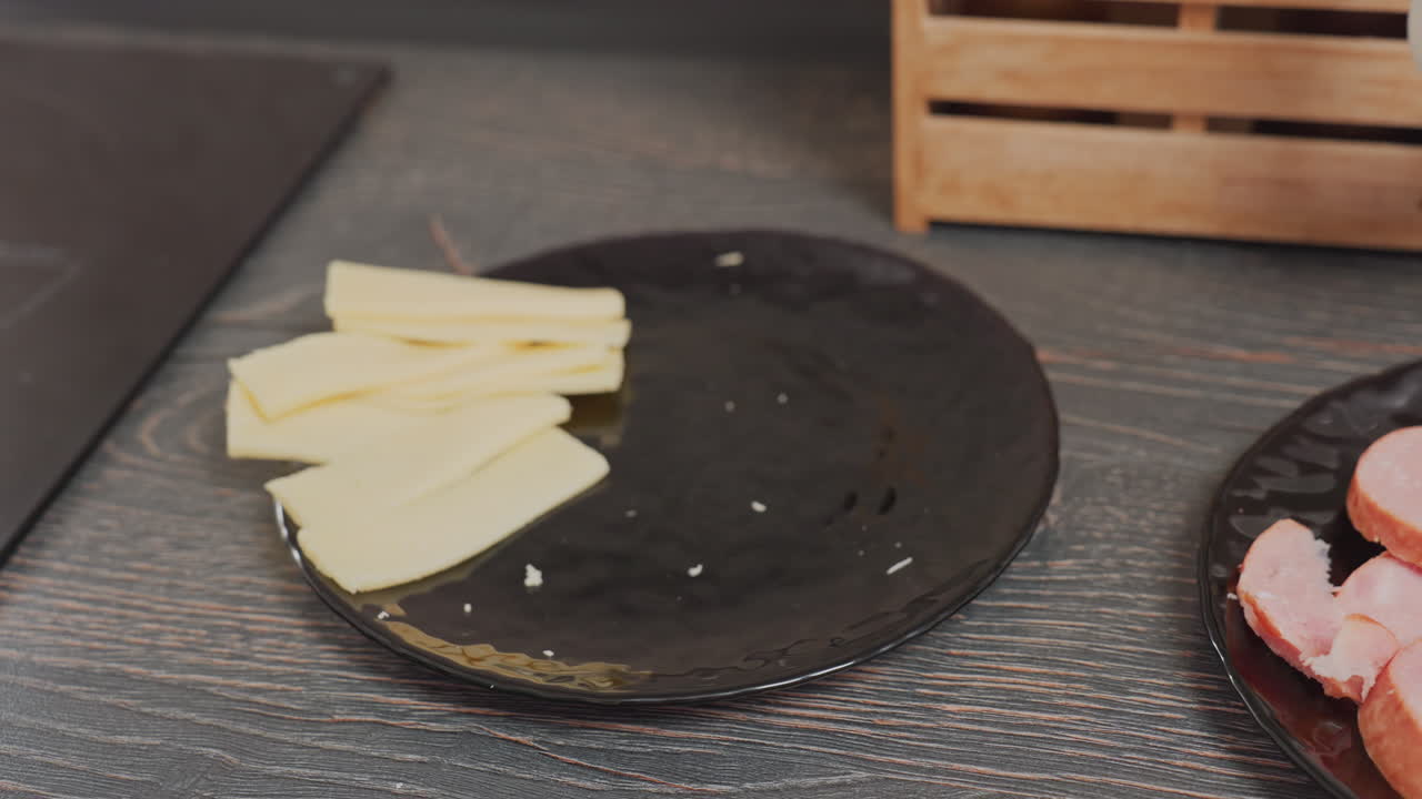 several slices of yellow cheese placed on black plate beside sliced sausage on matching plate sitting on dark wooden kitchen counter near spice rack in soft natural light kitchen setting