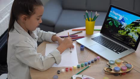 Una-Niña-Pequeña-Y-Seria-Estudia-Escritura-A-Mano-En-Línea-Usando-Una-Computadora-Portátil-En-Casa,-Una-Niña-Pequeña-Y-Feliz-Toma-Una-Lección-O-Clase-En-Internet