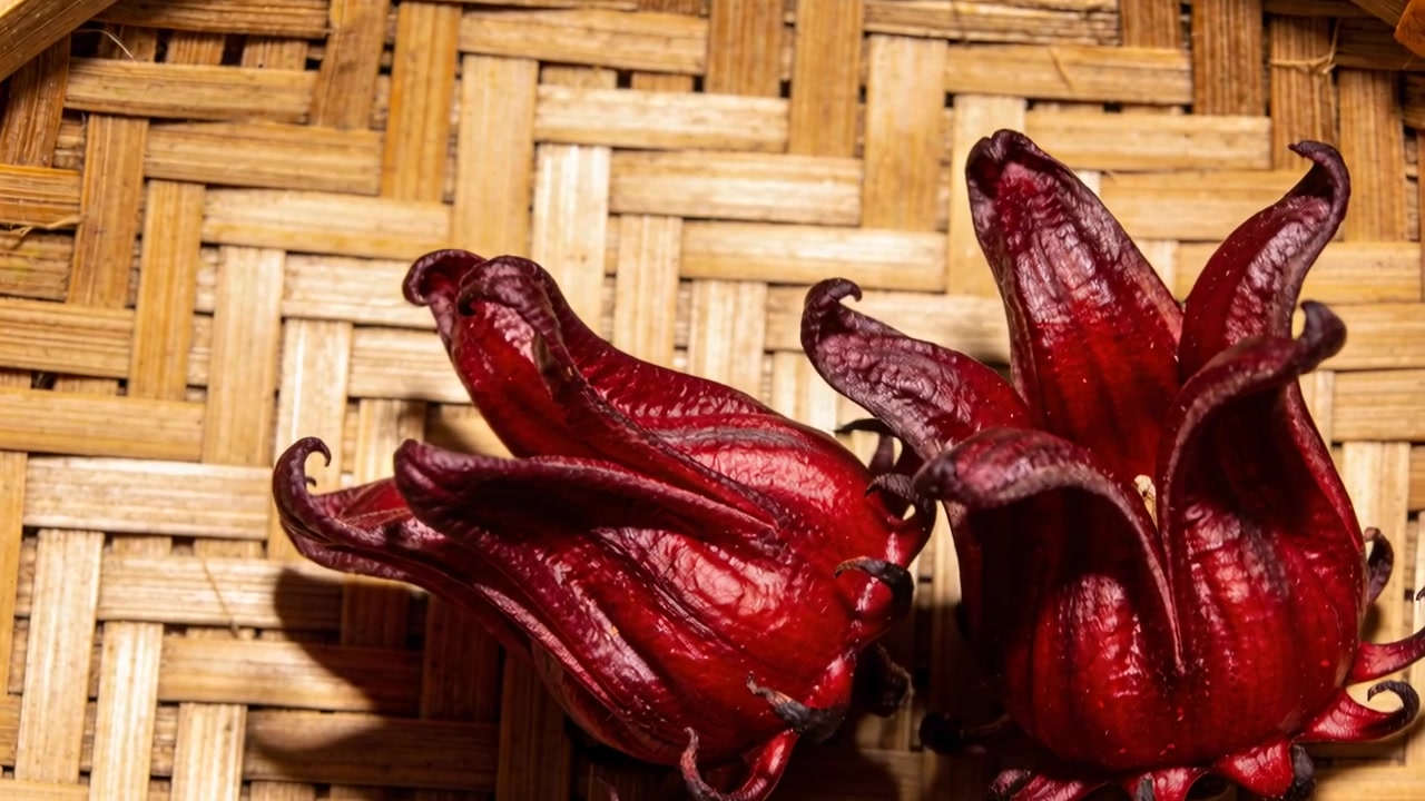 La telecamera si sposta rivelando due calici di roselle rosso intenso su un vassoio di bambù intrecciato, evidenziandone la trama.
