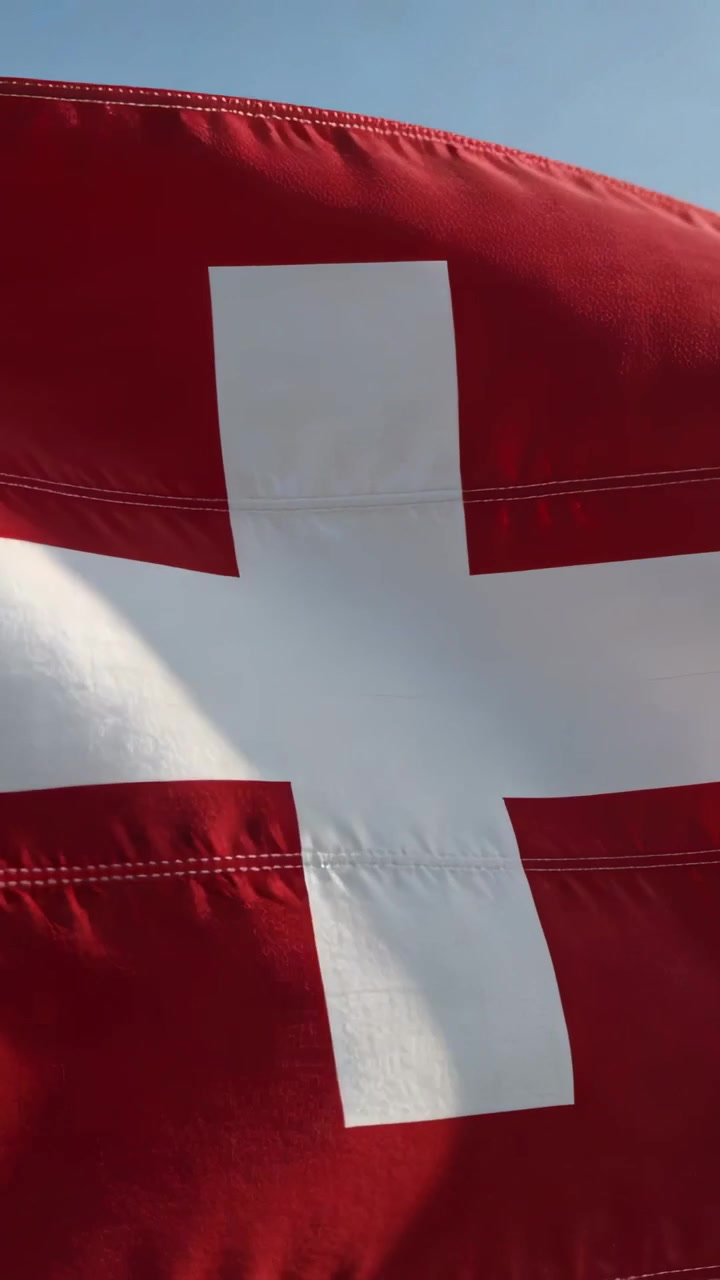 Vertikales Video: Wehende rote Flagge mit weißem Kreuz in der Mitte im Wind, Nähte leuchten im blauen Himmel.