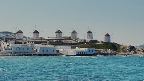 Stetige-Aufnahme-Der-Windmühlen-Von-Mykonos-An-Einem-Sonnigen-Tag-Mit-Meereswellen