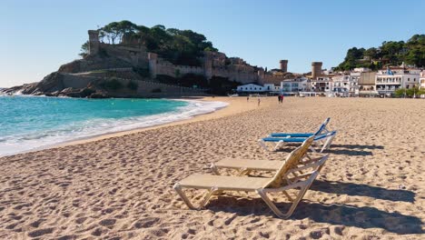 Deck-chairs-on-the-beach-with-the-background-of-walled-castle-over-the-sea-in-Tossa-de-Mar,-Girona-Spain-Costa-Brava-turquoise-water-beaches