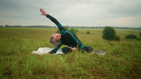 middle-aged woman lying on one side of yoga mat practicing side bend pose with arm extended, surrounded by nature in a vast grassy field under cloudy skies, with trees in the distance