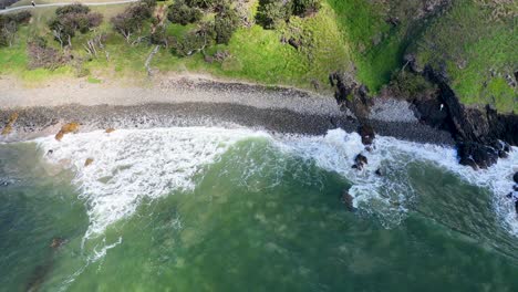 aerial footage captures waves crashing on byron bay's rocky shoreline, surrounded by lush greenery under bright daylight